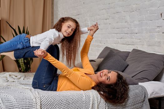 Love of young mother and daughter. They lie on the bed at home and have fun, they are fooling around, mother is holding her daughter on top of her legs