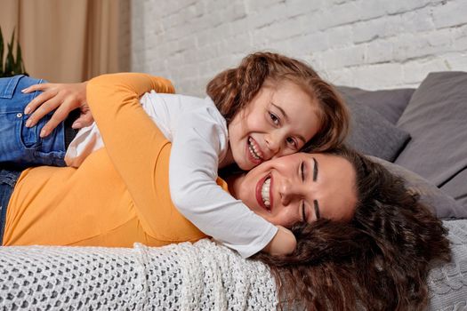 Love of young mother and daughter. They lie on the bed at home and have fun, daughter lies on top of mom, they hug each other