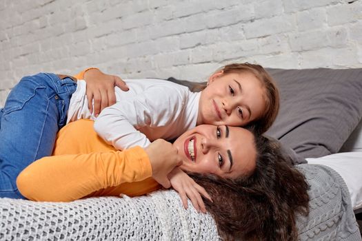 Love of young mother and daughter. They lie on the bed at home and have fun, daughter lies on top of mom, they hug each other