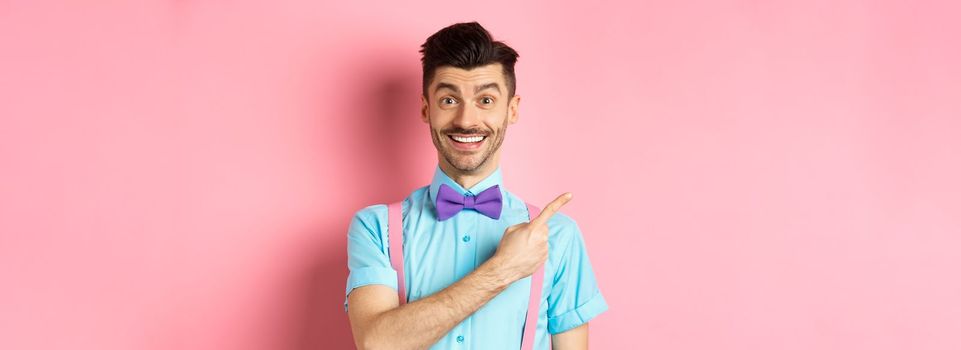 Portrait of handsome happy man showing logo, pointing left and smiling at camera, recommending shop offer, standing on pink background.