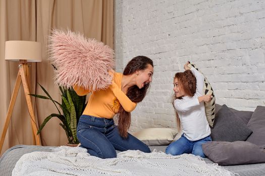 mom and daughter sit on bed, fooling around with pillow fights, they have fun, they looks happy