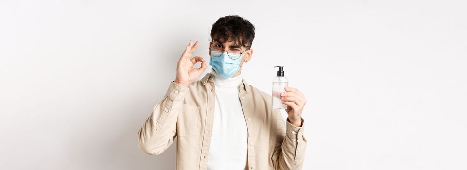 Health, covid and quarantine concept. Portrait of natural guy in glasses and face mask showing bottle of good hand sanitizer, make okay sign, recommending product, white background.