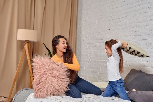 mom and daughter sit on bed, fooling around with pillow fights, they have fun, they looks happy