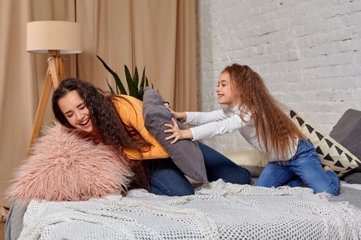 mom and daughter sit on bed, fooling around with pillow fights, they have fun, they looks happy