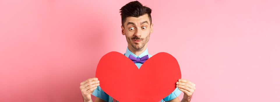 Valentines day concept. Cute boyfriend looking at big red heart, waiting for lover on romantic date, pink background.