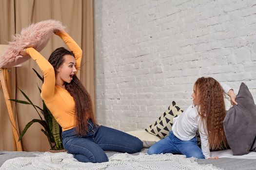 mom and daughter sit on bed, fooling around with pillow fights, they have fun, they looks happy