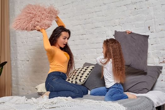 mom and daughter sit on bed, fooling around with pillow fights, they have fun, they looks happy