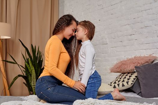 Love of young mother and daughter. They sit on the bed opposite each other and show emotions, touching their foreheads, and holding hands