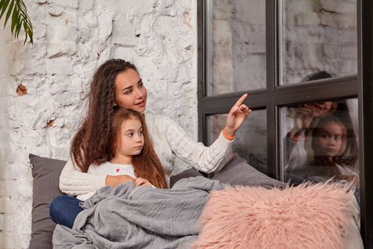 Mother and daughter sitting on sill near window in room. They show emotions and have fun