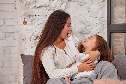 Mother and daughter sitting on sill near window in room. They show emotions and have fun