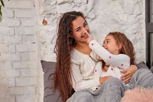 Mother and daughter sitting on sill near window in room. They show emotions and have fun