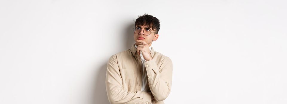 Handsome hipster guy in glasses look pensive at empty space, touching chin and frowning, stare up thoughtful, making choice with doubtful face, standing on white background.