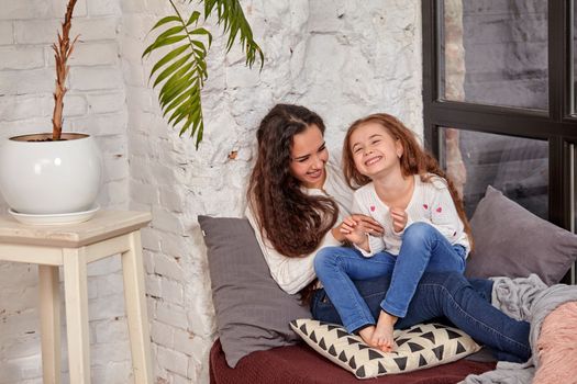 Mother and daughter sitting on sill near window in room. They show emotions and have fun