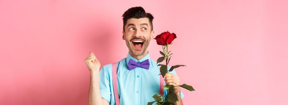 Romance and Valentines day concept. Cheerful man in bow-tie screaming from happiness, holding red rose and jumping on date, standing over pink background.