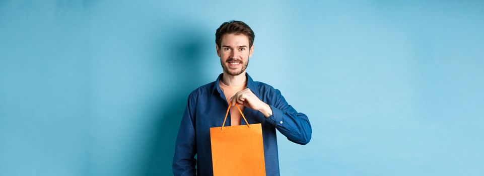 Handsome young man with beard, smiling and showing shopping bag, buying something in store, standing on blue background.