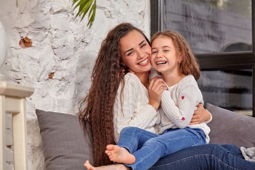 Mother and daughter sitting on sill near window in room. They show emotions and have fun