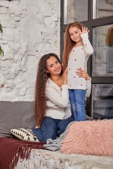 Mother and daughter sitting on sill near window in room. They show emotions and have fun