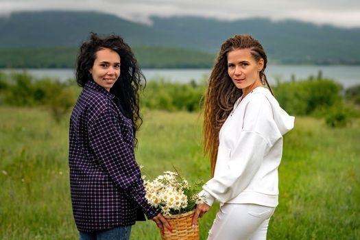 Two women enjoy nature in a field of daisies. Girlfriends hugging hold a bouquet of daisies and look at the camera