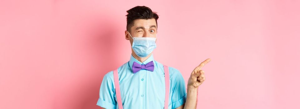 Covid-19, pandemic and health concept. Cheerful caucasian man in face mask winking at camera, showing good deal, pointing finger left at logo, pink background.