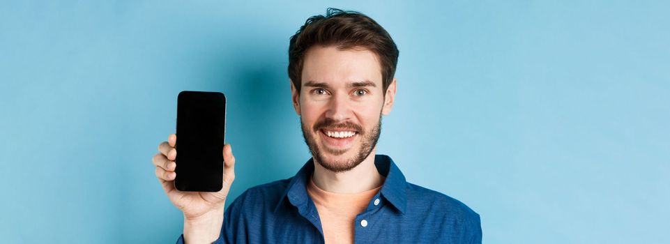 Close-up of smiling european man showing empty smartphone screen and demonstrating app, standing on blue background.