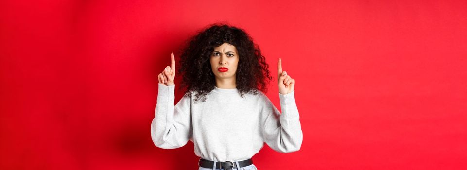 Grumpy young woman with curly hair frowning and grimacing unsatisfied, pointing fingers up at something bad, complaining on company, red background.