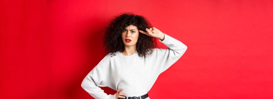 Are you stupid. Annoyed and confused girl with curly hair, pointing at head and scolding person being crazy or strange, standing on red background.