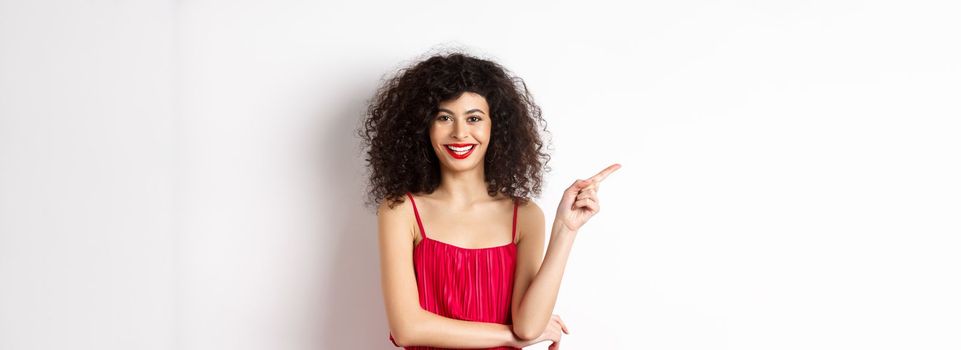 Beautiful happy woman with curly hairstyle, wearing festive dress, pointing finger left at logo and smiling, white background.