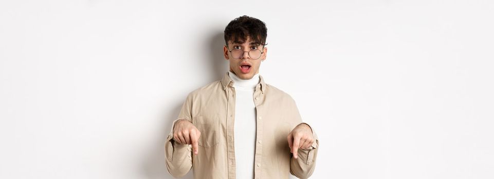 Shocked attractive young man showing something awesome, drop jaw and gasping in awe, pointing fingers down, standing on white background.