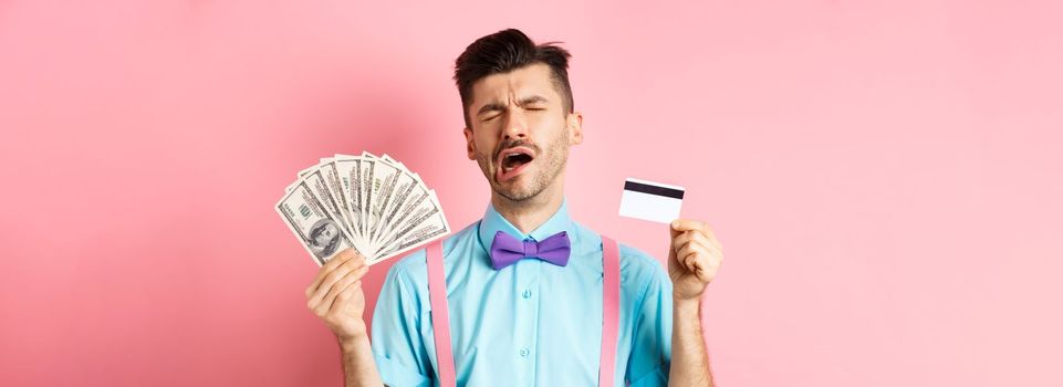 Sad crying man looking miserable, showing plastic credit card and money, standing upset on pink background.