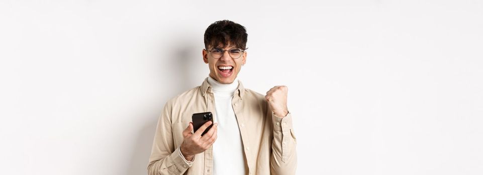 Technology and online shopping concept. Motivated and happy guy scream yes and show fist pump after winning on smartphone, achieve goal in app, white background.