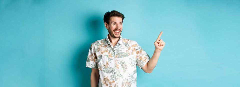 Excited smiling tourist pointing and looking aside at empty space, checking out promo offer, standing in hawaiian shirt on blue background.
