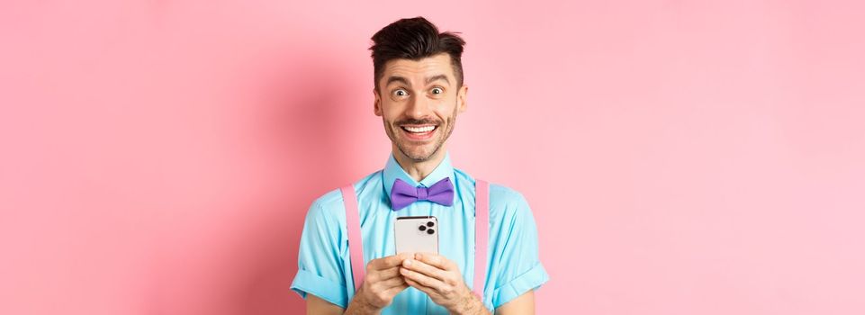 Online shopping. Happy guy in bow-tie, smiling at camera and using smartphone, standing on pink background.