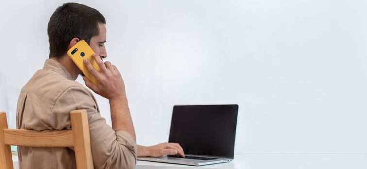 Young freelancer sitting at his work desk talking with smart phone call in front of his laptop. High quality photo with copy space for text banner.