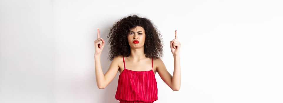 Disappointed frowning woman with makeup, wearing red dress on valentines, pointing fingers up and complaining, standing on white background.