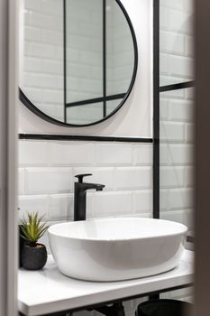 Bathroom with white brick walls, black faucet, an oval washbasin on a white table, a round mirror on a wall with black frames
