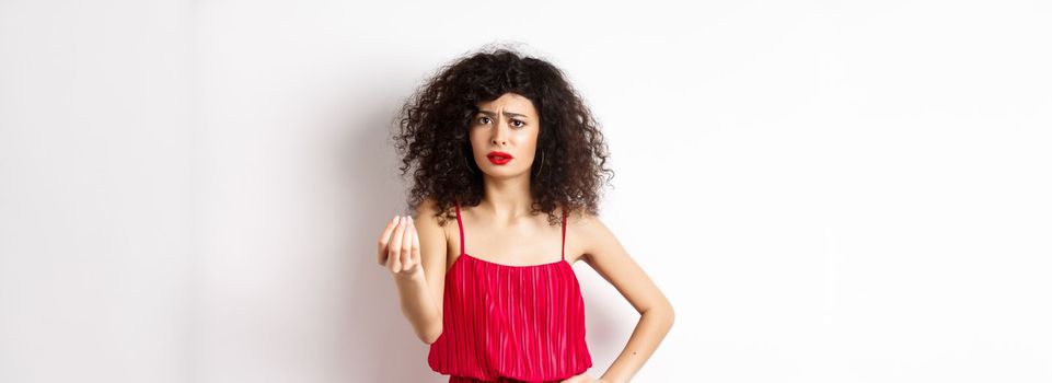 Confused woman with curly hair, wearing red dress, frowning and shaking hand, cant understand something, looking hesitant, standing on white background.