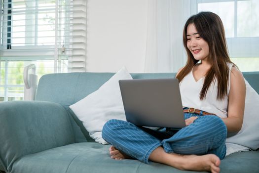 Happy woman typing email on notebook computer, Asian young female smiling sitting relaxing on sofa using laptop in living room at home, freelance browsing through the internet during free time