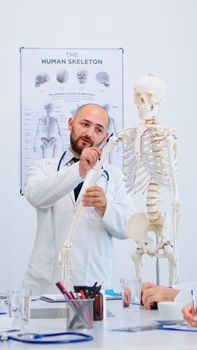 Medical doctor man pointing on cervical spine of human skeleton anatomical model, explaining to coworkers medical procedures in hospital meeting room. Physicians discussing about symptoms of disease