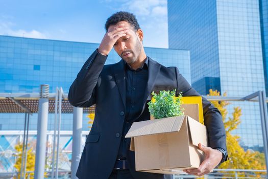 African American businessman fired with cardboard box with office supplies standing sad outside the building. High quality photo