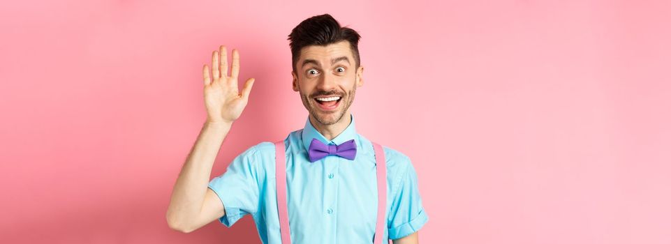 Friendly smiling man in funny bow-tie saying hello, waving hand to greet you, make hi gesture and looking happy yo see you, standing over pink background.
