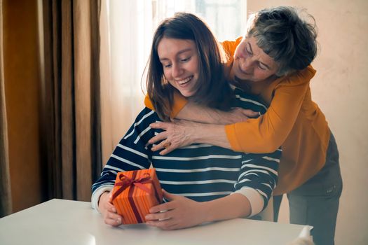 An elderly woman, a grandmother carefully hugs her granddaughter, gives her a surprise, makes a nice gift for the holidays, two women relatives have fun, a family happily spend time together.