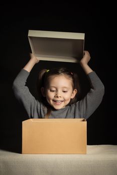 Emotional joyful little girl opens a long-awaited package with a gift, copy space on cardboard box, vertical.