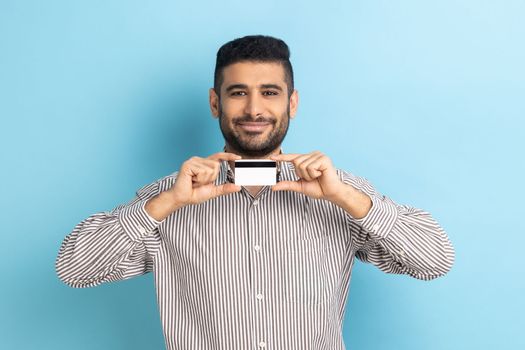 Businessman pointing finger at credit card, looking with happy expression, recommend bank or shopping discounts, wearing striped shirt. Indoor studio shot isolated on blue background.