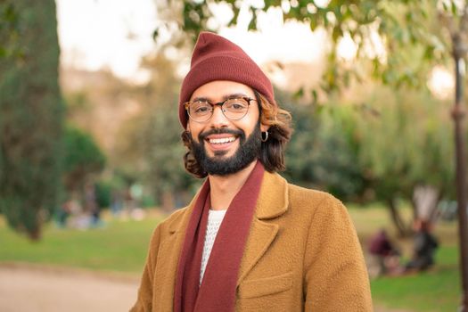 Portrait of a young adult man smiling looking at camera. Hipster man with glasses, beard, earrings, scarf and hat.