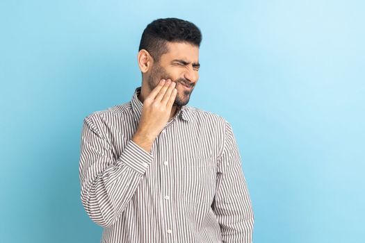 Unhappy bearded businessman feeling toothache, touching sore cheek, suffering from cavities, cracked teeth, gum recession, wearing striped shirt. Indoor studio shot isolated on blue background.