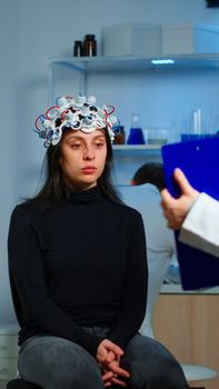 Medic researcher pointing at clipboard explaning treatment against brain disease to patient with eeg headset. Woman sitting in neurological scientific laboratory treating nervous system dysfunctions