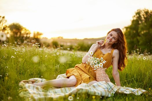 happy redhead woman sitting in a field of daisies on a plaid during sunset and smiling at the camera holding a flower in her hand. High quality photo