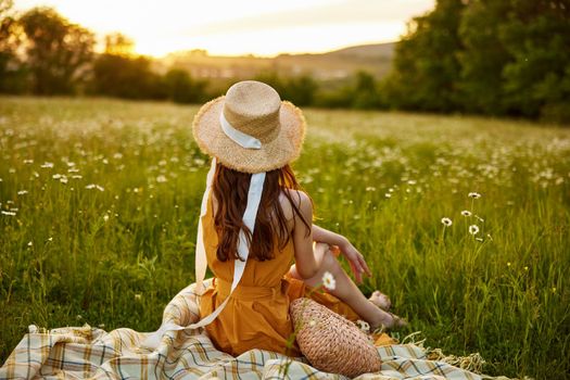 a woman in a wicker hat sits with her back to the camera in a chamomile field and looks at the sunset. High quality photo