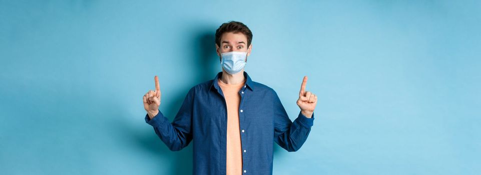 Happy guy in medical mask feeling excited about good news, pointing fingers up and smiling, standing on blue background.