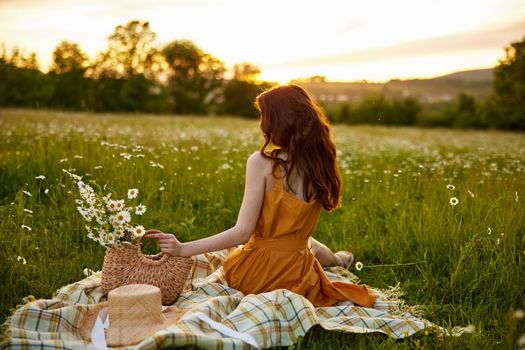 a woman with beautiful, long well-groomed hair sits in an orange dress in a chamomile field and looks at the sunset. High quality photo